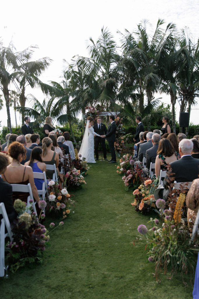 image displaying the aisle of the wedding ceremony with the bride and groom standing at the arch holding hands and all their guests are seated. The florals depicted lining the aisle are jewel tones, green, mauve, deep maroon, red and pink, portraying a moody and romantic design. The chairs set up for the ceremony are white folding chairs facing a floral arch with palm trees. the aisle and ceremony is set on green grass at the Carlsbad Cape Rey Hotel in San diego. The ceremony is facing the coast at the beach. 