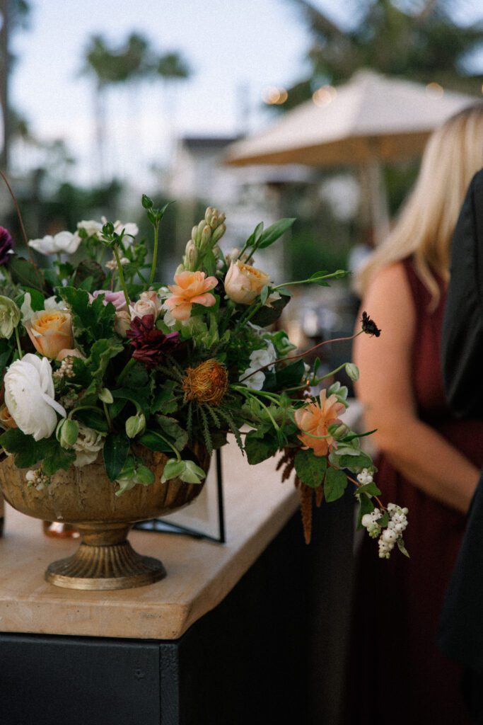 images depicts a floral arrangement on top of a black wood bar with a light wood table top. The floral arrangement has orange, white and maroon florals amongst lush greenery. the arrangement is sitting in a antique gold footed bowl. The arrangement ties into the moody and romantic theme of the wedding