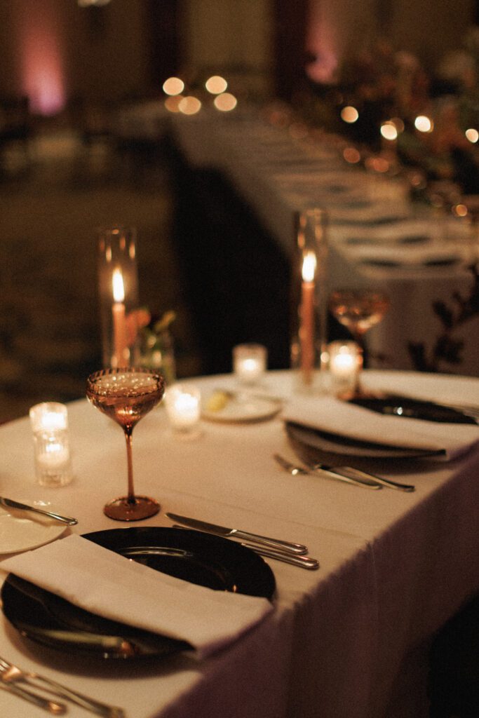 the image depicts the table top decor for the sweetheart table from a different view. this includes a black charger plate, white napkin, silver flatware, a dusty rose water goblet . the tables is a half moon. candles are sprinkled throughout the table displays as well, create the perfect moody and romantic ambience. 
