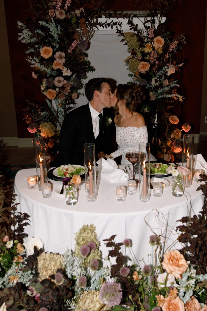 the image depicts the bride and groom sitting at their sweetheart table that is a half moon shape with a white linen. the table is covered in taper candles and tea light candles. on the table is also place settings with black charger plates, and other flatware for dinner. in front of the table on the floor, the aisle arrangements from ceremony have been moved here creating a beautiful moody romantic display for the couple. the aisle arch has also been placed behind the sweetheart table to add a backdrop for the couple as well. the couple is sitting in their seats kissing for a photo.