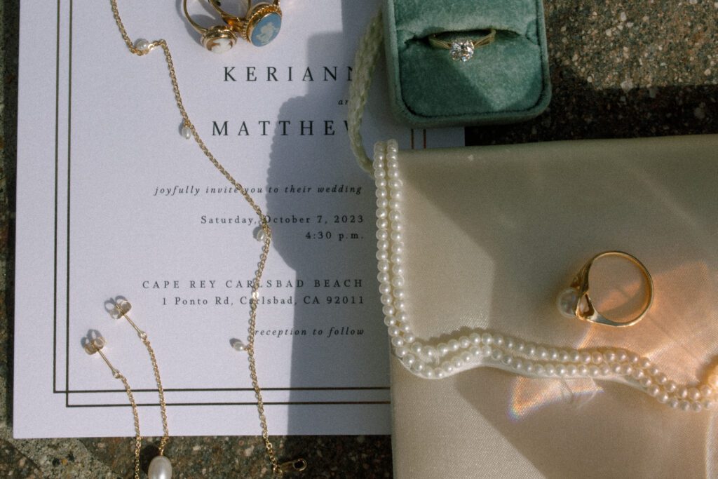 image displaying an assortment of wedding items such as wedding invitation, wedding rings, engagement ring inside a green velvet ring box, pearl beaded white purse, gold necklace with small pearls and matching gold earrings with pearls