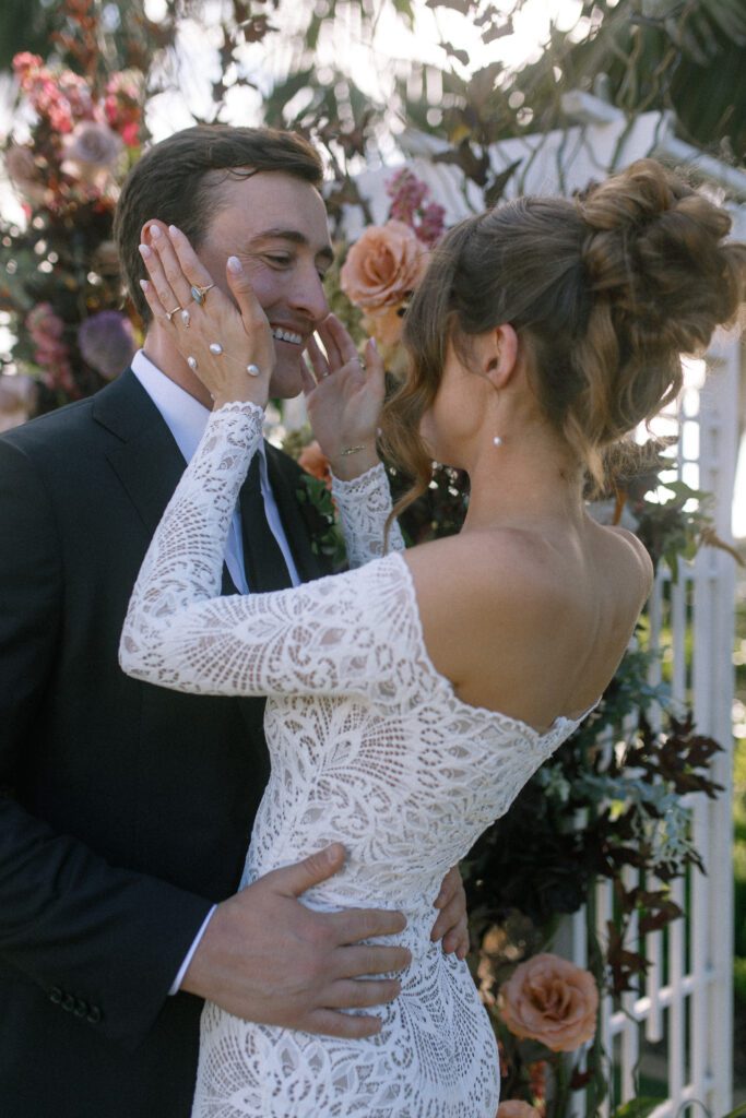 image displaying the bride and groom smiling at each other after they have just been married. The bride is holding the grooms face, pulling him in for a kiss. They are standing near their floral arch from their ceremony. Their wedding was a moody, romantic wedding at cape rey carlsbad in san diego