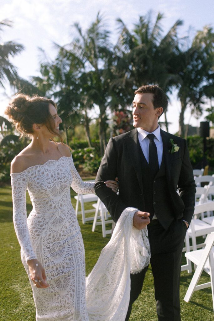 image displaying the bride and groom smiling and walking together. The groom is holding the brides dress casually as they walk together. Their wedding was a moody, romantic wedding at cape rey carlsbad in san diego