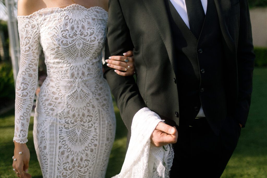 the bride and groom stand side by side to show their outfits/attire. the image depicts only from their shoulders to their knees. Their arms are interlocked and the groom is holding the end of the brides dress around his arm. the brides dress is an intricate lace dress that is form fitting. The dress is off the shoulder and has long lace sleeves. The groom is wearing a three piece black suck with a white undershirt and a plain black tie. 