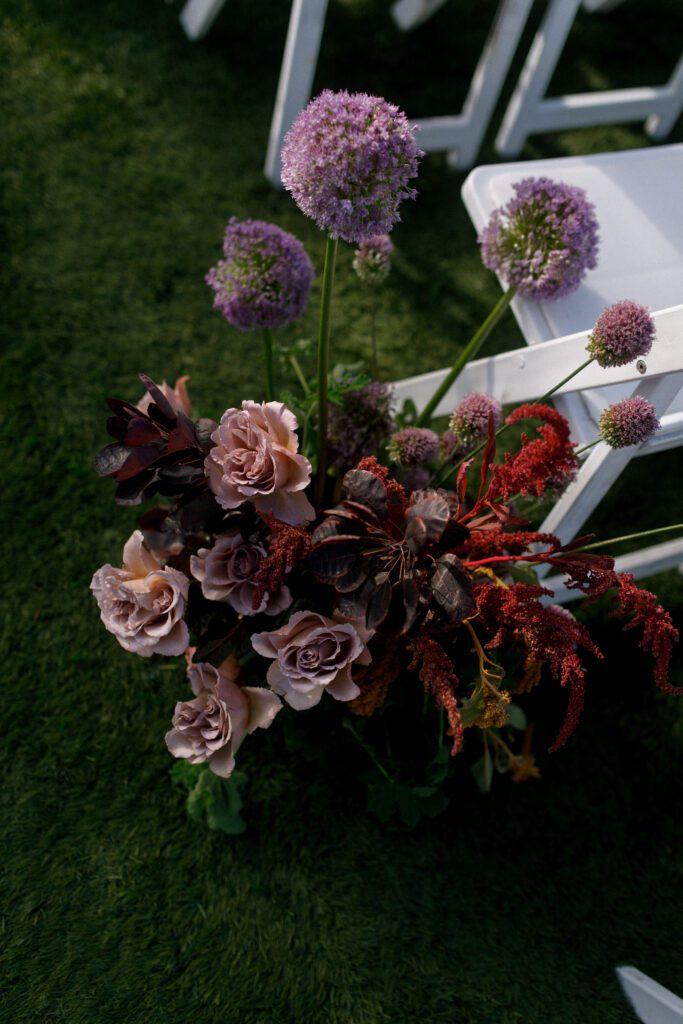the image depicts a floral arrangement up close from the aisle of the wedding ceremony. the florals are purple, pinks, maroon and red with lush green florals. the flowers have a whimsical and natural style and create a moody and romantic design and vibe to the wedding.