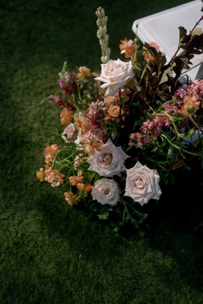 the image depicts a floral arrangement up close from the aisle of the wedding ceremony. the florals are purple, pinks, maroon and orange with lush green florals. the flowers have a whimsical and natural style and create a moody and romantic design and vibe to the wedding.