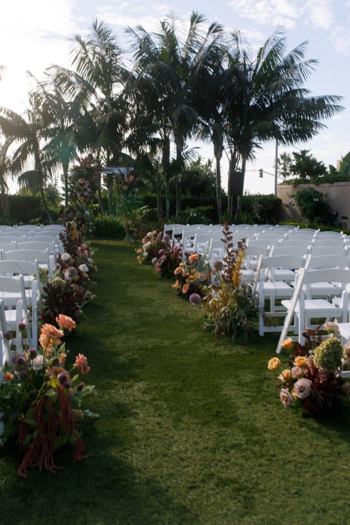 image displaying the aisle of the wedding ceremony before guest have arrived. The florals depicted are jewel tones, green, mauve, deep maroon, red and pink, portraying a moody and romantic design. The chairs set up for the ceremony are white folding chairs facing a floral arch with palm trees. the aisle and ceremony is set on green grass at the Carlsbad Cape Rey Hotel in San diego. The ceremony is facing the coast at the beach. 