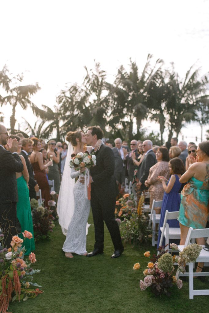 image displaying the aisle of the wedding ceremony with the bride and groom standing halfway down the aisle after they have been married. They stopped to share a kiss. Their guests are standing around them, clapping and cheering in their places. The florals depicted lining the aisle and in the bouquet are jewel tones, green, mauve, deep maroon, red and pink, portraying a moody and romantic design. The chairs set up for the ceremony are white folding chairs facing a floral arch with palm trees. the aisle and ceremony is set on green grass at the Carlsbad Cape Rey Hotel in San diego. The ceremony is facing the coast at the beach. 