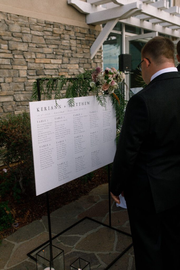 image depicts a seating chart at the cocktail hour of the wedding. a guest is standing near it reading it. the sign is horizontal and configured on a black metal stand with a floral arrangement at the top right corner of the seating chart. the floral arrangement has lush greenery, maroon, pink and white flowers. 