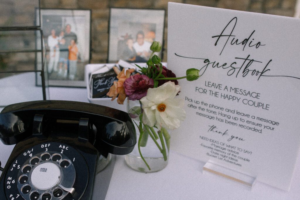 image depicts the guestbook available for the guests at the wedding. the guest book is an old fashion rotary phone in black that has a sign beside it giving instructions. the instructions tell the guests to pick up the phone and speak a message into the phone for the couple, whatever message they say will be saved and recorded for the couple to listen to after the wedding. next to the phone is a small bud vase with a couple florals inside, matching the theme of the other florals of the wedding. behind the bud vase is a bowl of custom match boxes set out as a souvenir for the guests to take home. there are a couple photos in the background in frames on the table as well. on the gift table with thee items is also a black metal and glass see through card box.