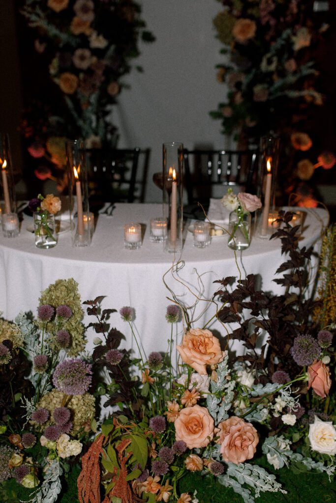 the image depicts the bride and grooms sweetheart table that is a half moon shape with a white linen. the table is covered in taper candles and tea light candles. on the table is also place settings with black charger plates, and other flatware for dinner. in front of the table on the floor, the aisle arrangements from ceremony have been moved here creating a beautiful moody romantic display for the couple. the aisle arch has also been placed behind the sweetheart table to add a backdrop for the couple as well. the bride and groom are not seating, it is just a photo of the decor and table