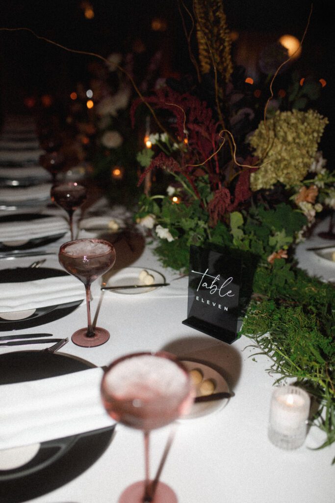 the image depicts the table top decor for the guest tables. this includes a black charger plate, white napkin, silver flatware, a dusty rose water goblet and lush floral centerpieces lining the center of the tables. the tables are long banquet tables. the table numbers are black acrylic with white writing. candles are sprinkled throughout the table displays as well, create the perfect moody and romantic ambience. offering a different view of the table, close up