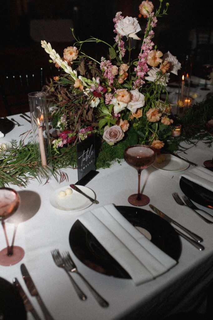 the image depicts the table top decor for the guest tables. this includes a black charger plate, white napkin, silver flatware, a dusty rose water goblet and lush floral centerpieces lining the center of the tables. the tables are long banquet tables. the table numbers are black acrylic with white writing. candles are sprinkled throughout the table displays as well, create the perfect moody and romantic ambience. offering a different view of the table