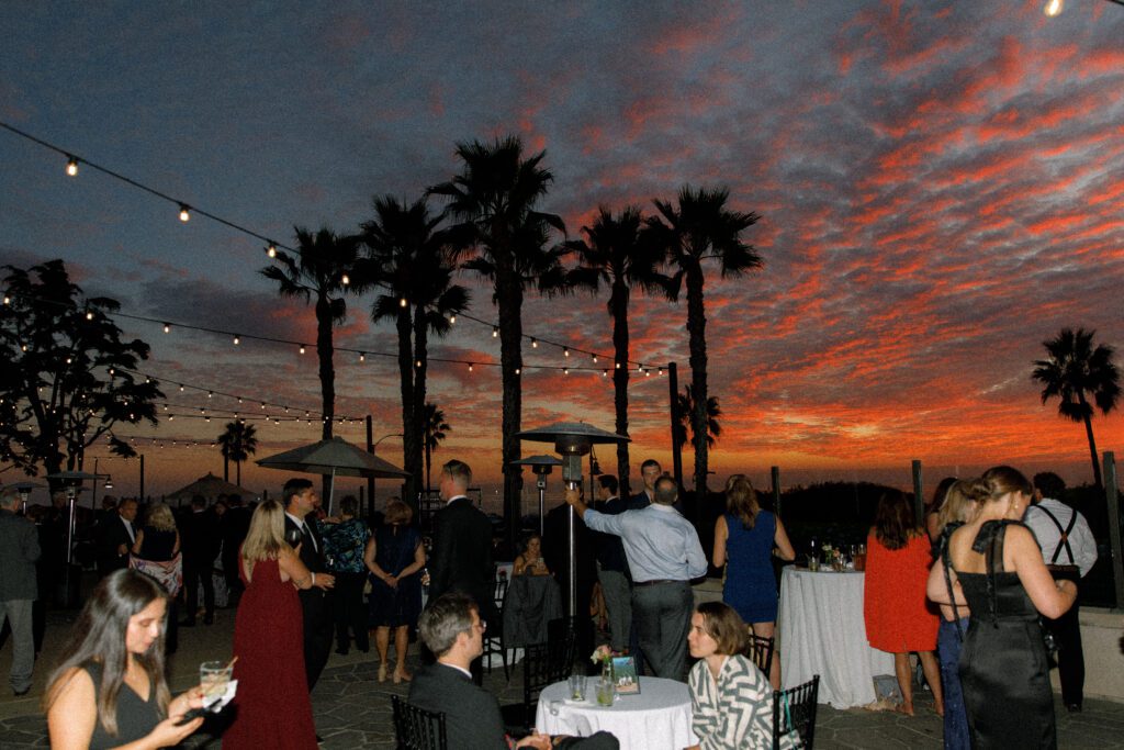 the image depicts the sunset during cocktail hours. the cocktail hour takes place on an outdoor terrace/patio next to the beach in san diego, california. the guests are mingling, some standing and some sitting at a cocktail table conversing. the sunset is purple, orange, blue, red, pink and yellow.