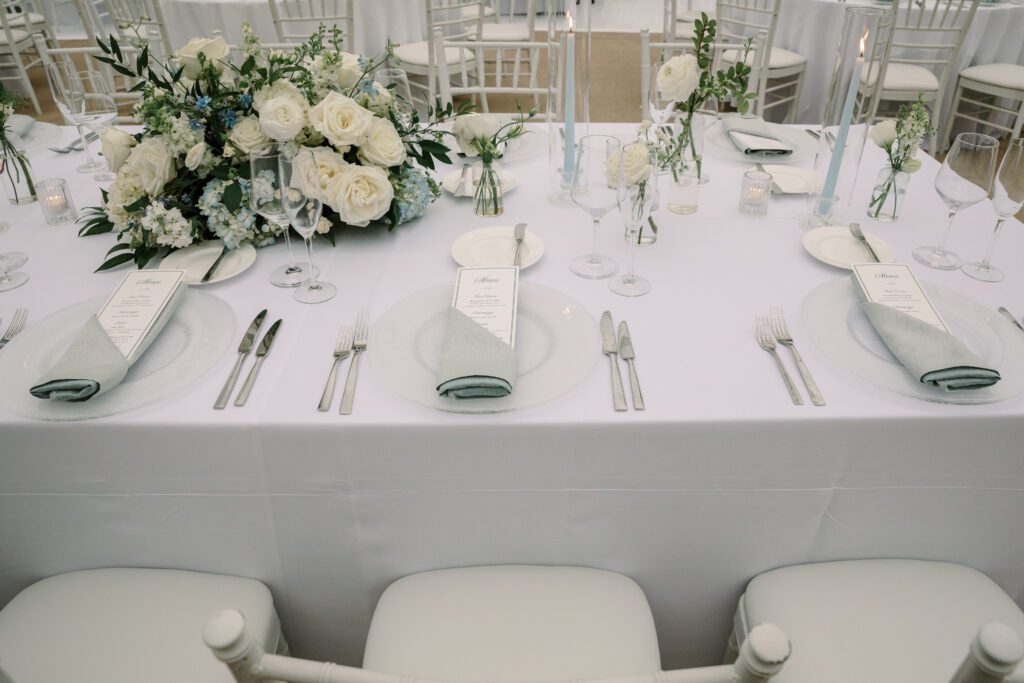 a close up of a place setting on the dinner reception tables. the floral arrangement is blue and white and green. the charger place is a clear glass with a gray blue napkin on top folded and holding the custom printed menu. the silverware is silver and there are classic glassware