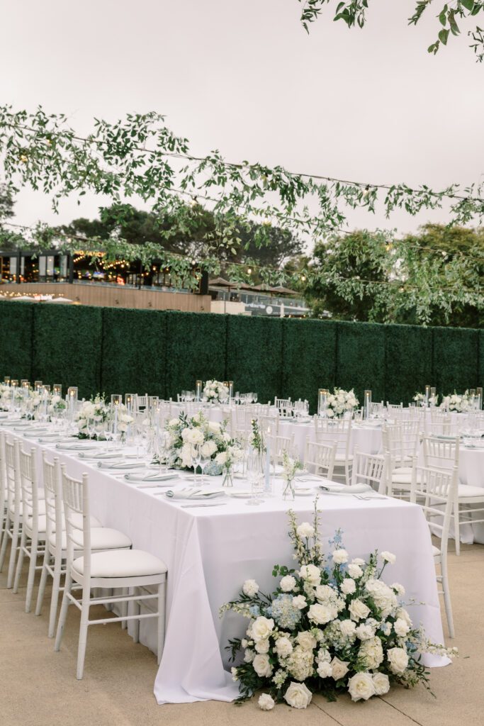 the dinner reception tables are long banquet tables and have green and white floral arrangements on them. the linens are white and the chairs are white. creating a coastal and timeless theme