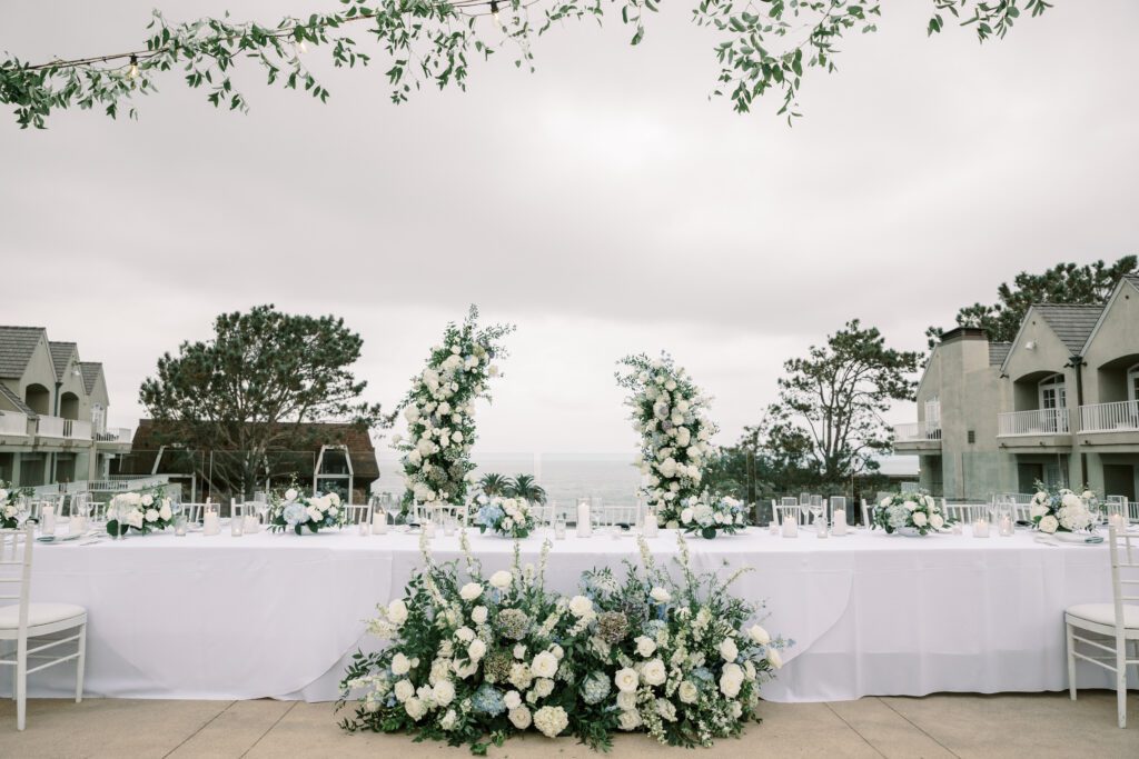 the head table at the dinner reception. the table is a long banquet table with a white linen. the chairs surrounding are white and the floral arrangements on top and on the floor are green and white with light blue accents. the ceremony arch is being repurposed to frame the couple behind their seats, creating a coastal and timeless theme