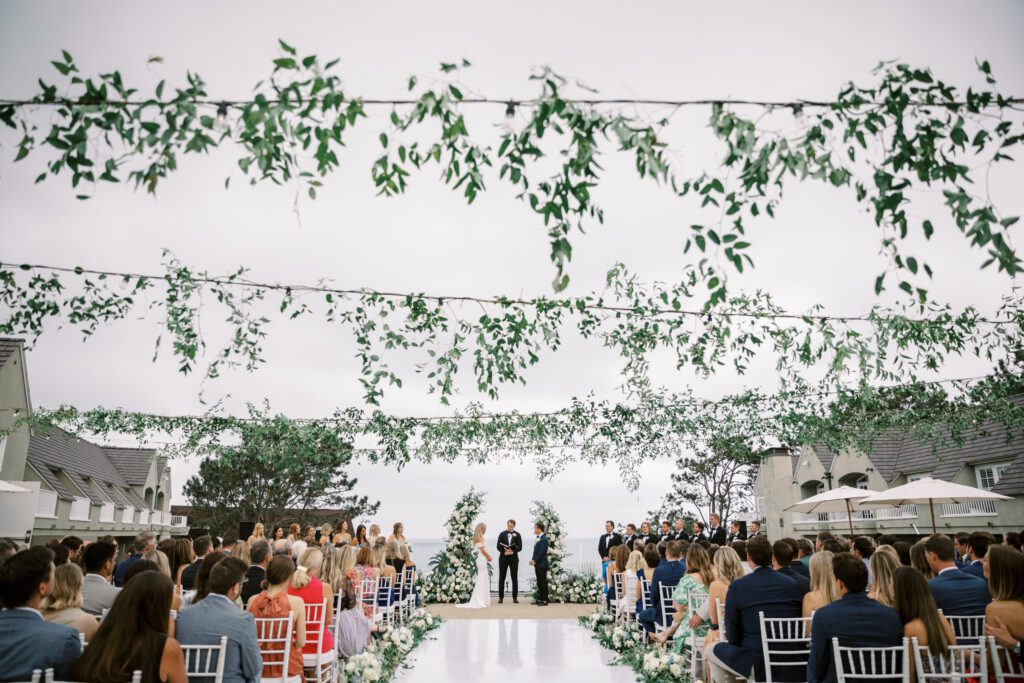 depicts a wedding ceremony taking place. the bride and groom are standing in front of the floral arch. the  guests are seated in their chairs. the aisle is a white vinyl material and lined with flowers and candles. there is greenery hanging above on the market lights. the ceremony is facing/ overlooking the ocean.creating a coastal and timeless theme