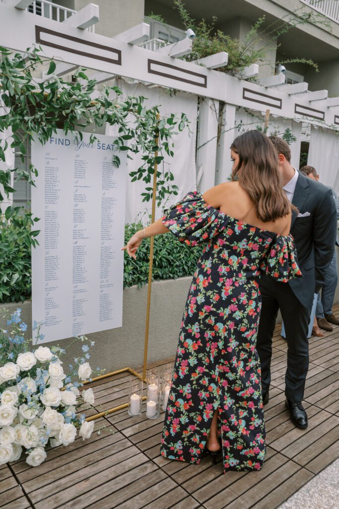 floral arrangement on the floor in front of the seating chart. the flowers are white roses and light blue accents.  the seating chart is white with blue writing. the stand is gold and has greenery framing the seating chart.creating a coastal and timeless theme