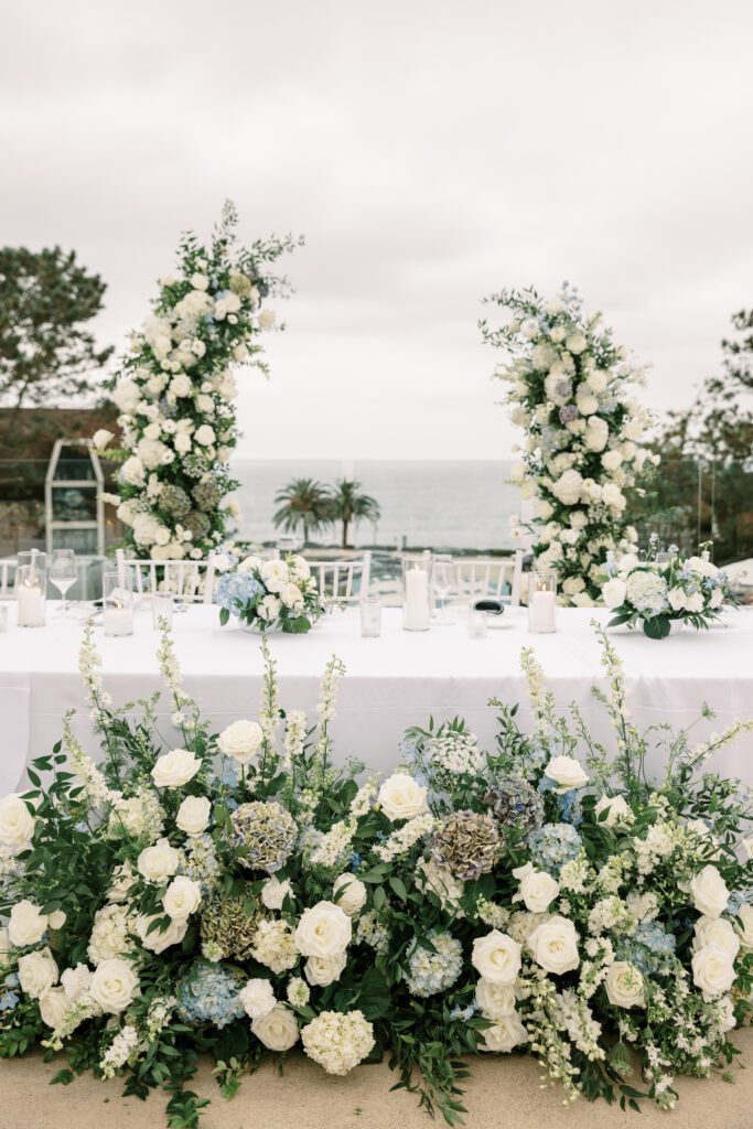 the head table at the dinner reception. the table is a long banquet table with a white linen. the chairs surrounding are white and the floral arrangements on top and on the floor are green and white with light blue accents. the ceremony arch is being repurposed to frame the couple behind their seats.creating a coastal and timeless theme