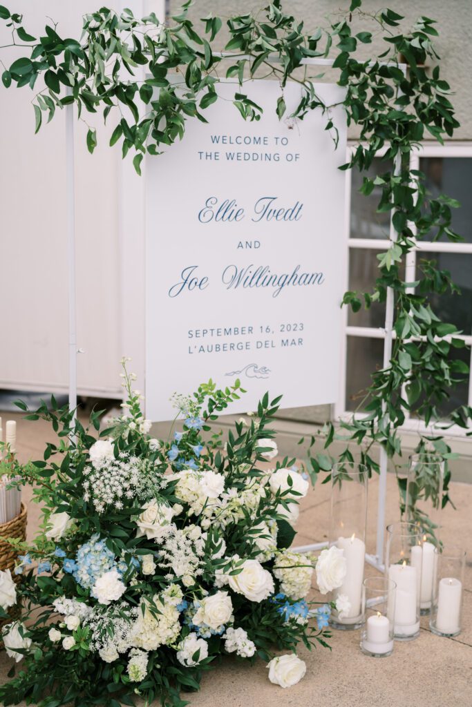 depicts a welcome sign at a wedding that reads " Welcome to the wedding of ellie and joe" The sign is white with delicate light blue writing. The sign is surrounded by lush greenery and a floral arrangement on the floor in front. the florals are white and blue and ivory white greenery. on the floor is also tall candles.creating a coastal and timeless theme