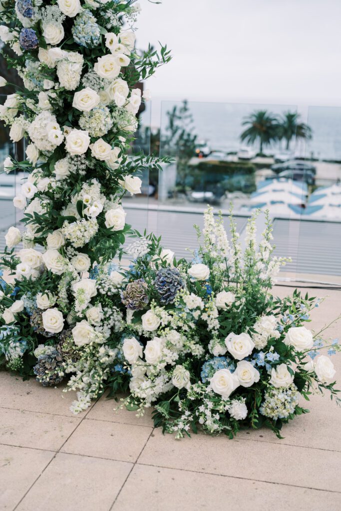 depicts a lush floral arrangement for the wedding. the arrangement is part of the arch for the ceremony. The florals are light blue and white with greenery filled in around the flowers.creating a coastal and timeless theme