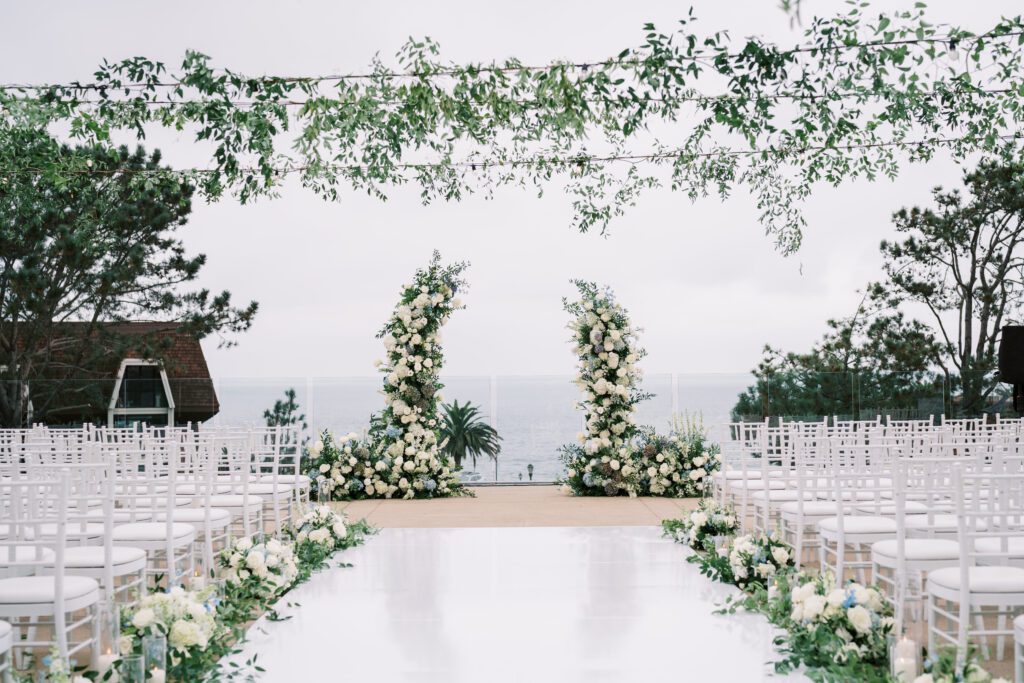 depicts a wedding ceremony before it has taken place. the guests are not yet seated in their chairs. the aisle is a white vinyl material and lined with flowers and candles. there is greenery hanging above on the market lights. the ceremony is facing/ overlooking the ocean.creating a coastal and timeless theme