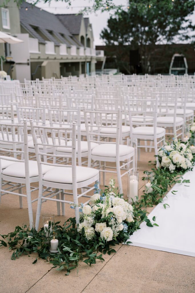 depicts the floral arrangments decorating the ceremony aisle. the aisle is vinyl white and has greenery and flowers in light blue and white along the edges. the chairs are white ad facing the ocean.