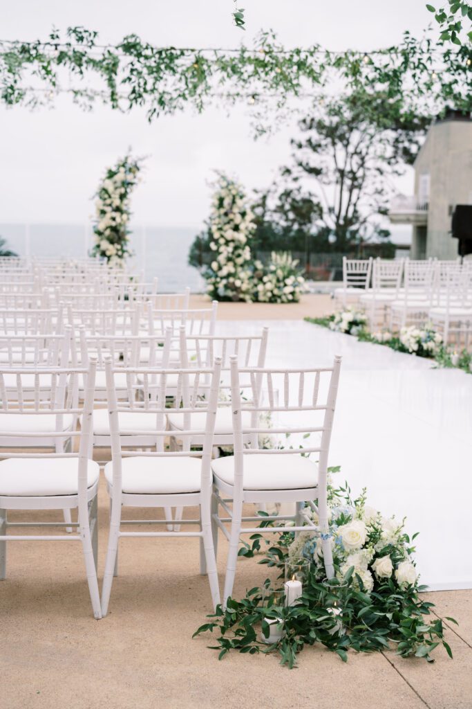 depicts a wedding ceremony before it has taken place. the guests are not yet seated in their chairs. the aisle is a white vinyl material and lined with flowers and candles. there is greenery hanging above on the market lights. the ceremony is facing/ overlooking the ocean. a different angle.creating a coastal and timeless theme