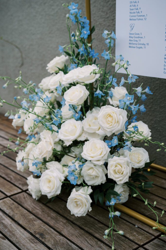 floral arrangement on the floor in front of the seating chart. the flowers are white roses and light blue accents. 