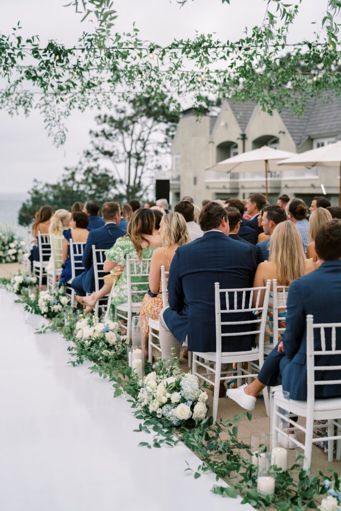 guests are seated for a wedding ceremony. the couple has not come down the aisle yet. the aisle is white and lined with florals and candles.creating a coastal and timeless theme