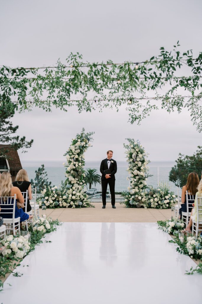guests are seated for a wedding ceremony. the couple has not come down the aisle yet. the aisle is white and lined with florals and candles. the officant is standing under the aisle waiting for the couple.creating a coastal and timeless theme