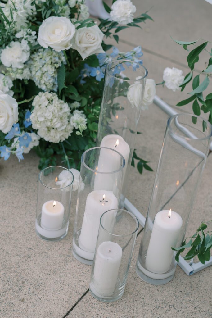 depicts a close up of the candles decorating the ceremony aisle. they are tall pillar candles inside a glass holder of varying heights.they are situated next to a floral arrangement