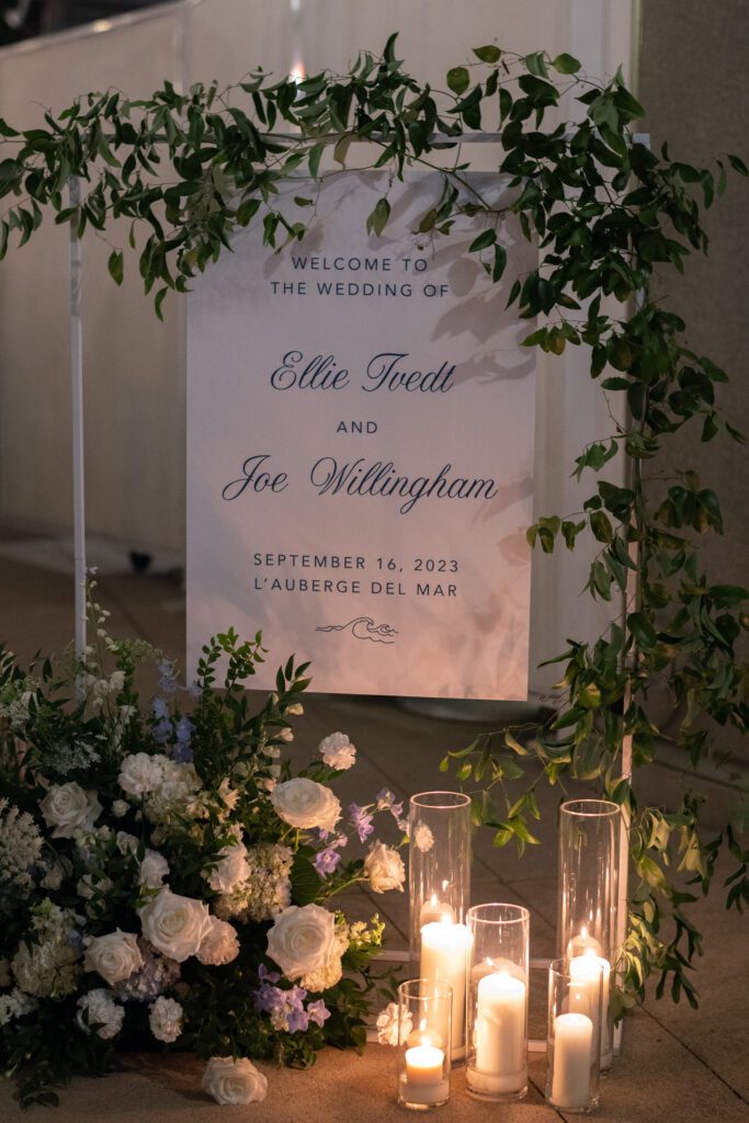 depicts a welcome sign at a wedding that reads " Welcome to the wedding of ellie and joe" The sign is white with delicate light blue writing. The sign is surrounded by lush greenery and a floral arrangement on the floor in front. the florals are white and blue and ivory white greenery. on the floor is also tall candles.creating a coastal and timeless theme. it is night time and the candles are illuminating the sign