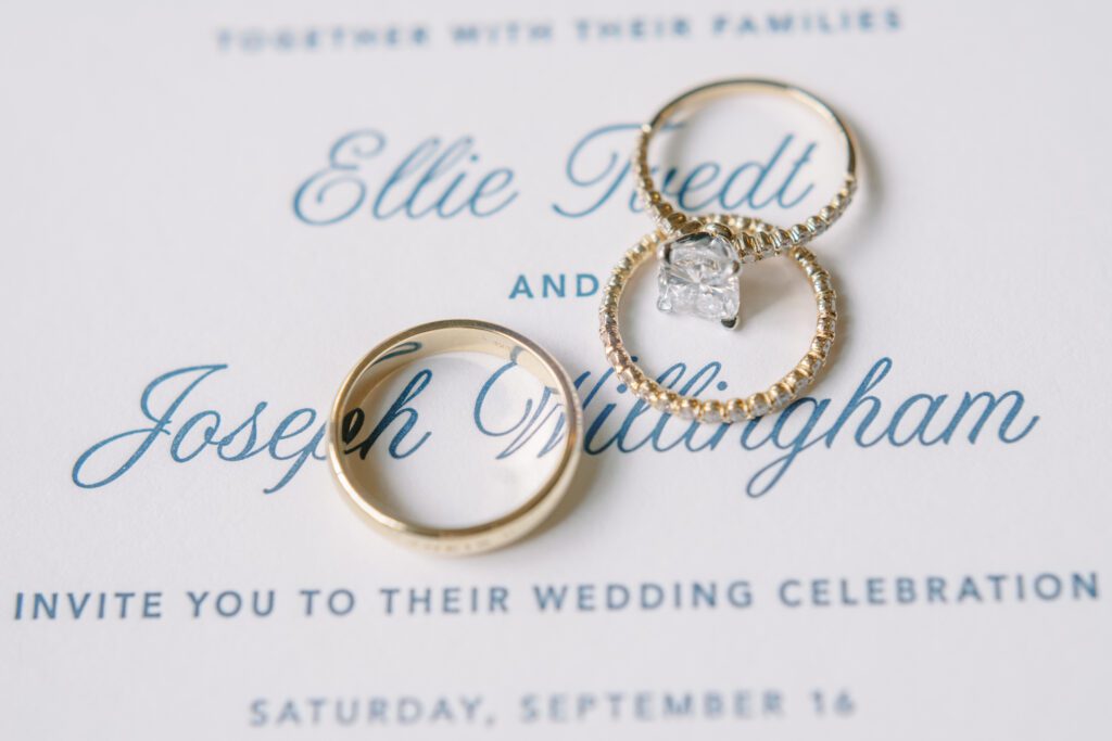 depicts a wedding invitation that is white with delicate blue writing. There are 3 gold wedding rings on top, the photo is up close to capture the details.