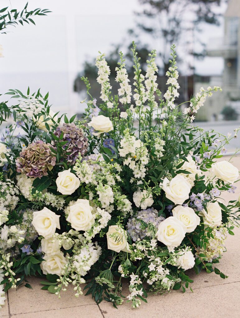 depicts a lush floral arrangement for the wedding. The florals are light blue and white with greenery filled in around the flowers and some accents of purple flowers.