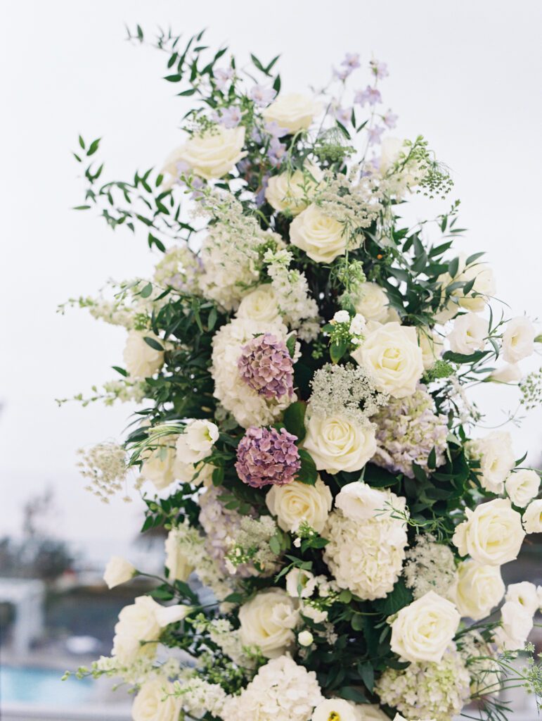 depicts a lush floral arrangement for the wedding. The florals are light blue and white with greenery filled in around the flowers and some accents of purple flowers. The arrangement ispart of the ceremony arch.