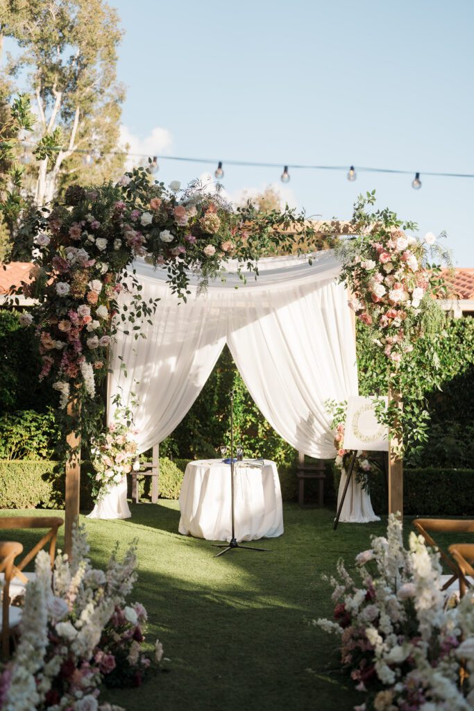 a wedding ceremony is taking place  in a garden courtyard. the decor and set up is white and pinks with greenery and light wood accents