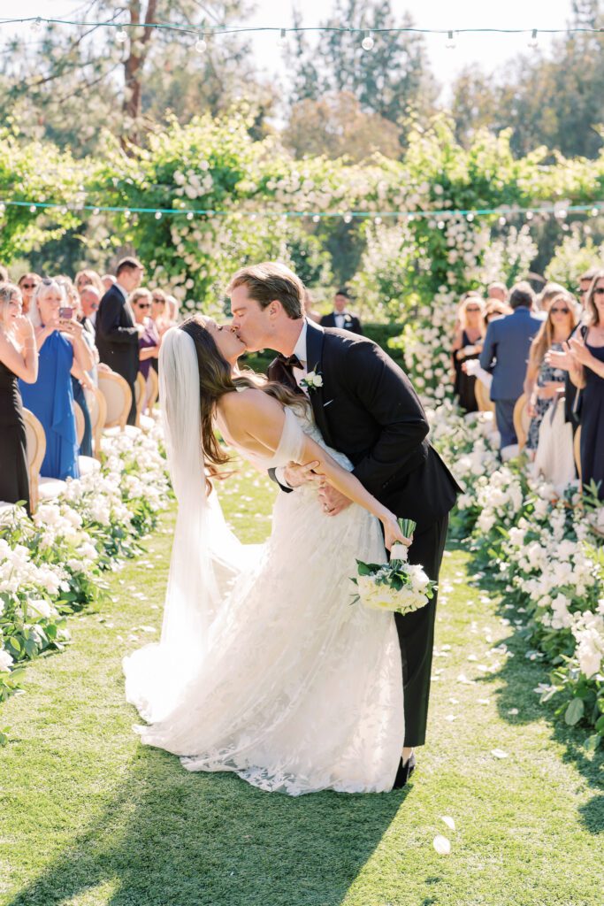 bride and groom kiss at their wedding ceremony. grooms dips the bride. they are surrounded by white florals