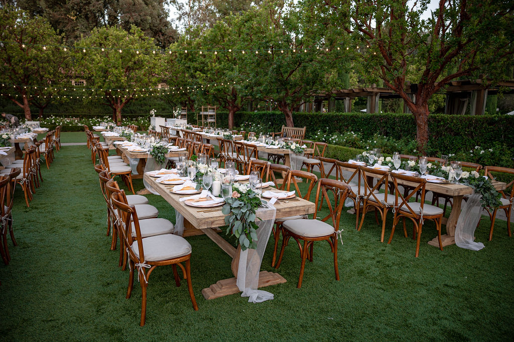 a wedding reception is taking place at rancho bernardo inn on the aragon lawn outdoors. the decor is wood tables and chairs with greenery