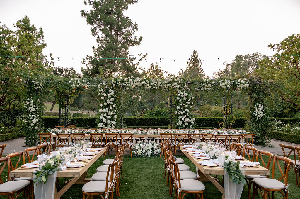 a wedding reception dinner is decorated and set up outdoors on a grassy courtyard  garden surrounded by white florals