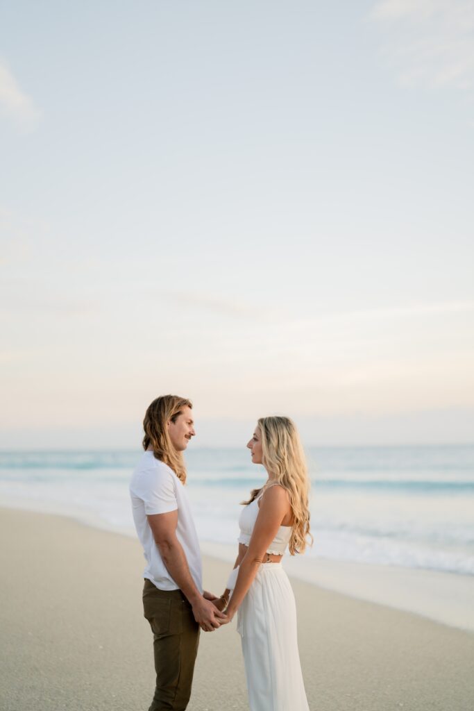 Newly engaged couple at the beach holding hands and looking at each other and demonstrating our engagement photo tips and styling advice