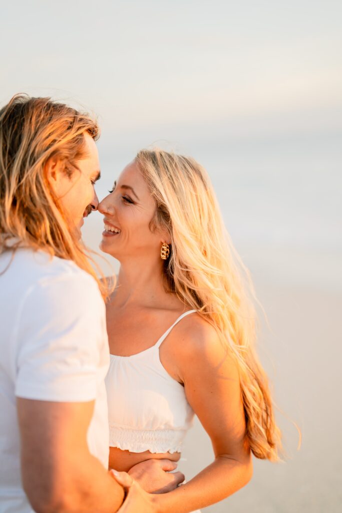 Newly engaged couple at the beach looking at each other smiling and demonstrating our engagement photo tips and styling advice