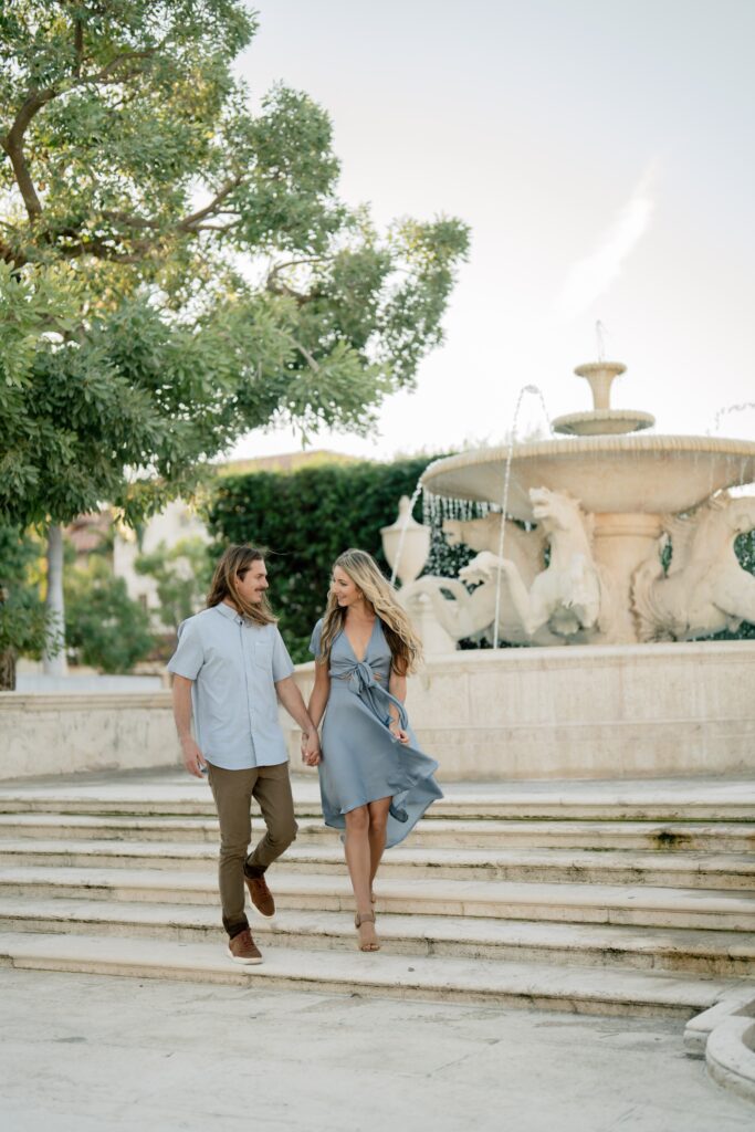 Newly engaged couple at a park with a fountain holding hands and walking together and demonstrating our engagement photo tips and styling advice